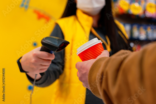 A girl seller in a medical mask from a virus holds a barcode scanner in her hands. A man with a glass of coffee in his hand pays at the checkout.