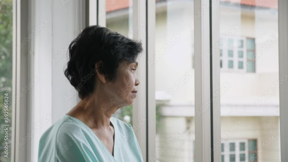 Indoors, close up, side view portrait of worried senior Asian woman wear green patient outfit, stand near the windows, look outside sadly Indoors, close up, side view portrait of worried senior Asian woman wear green patient outfit, stand near the windows, look outside sadly