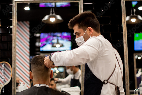 The young and elegant barber with mask cuts the hair of a client with Caucasian features