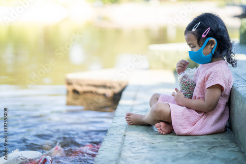 Fototapeta Naklejka Na Ścianę i Meble -  Cute little girl sitting at pond wearing protective face mask feeding carp fishes