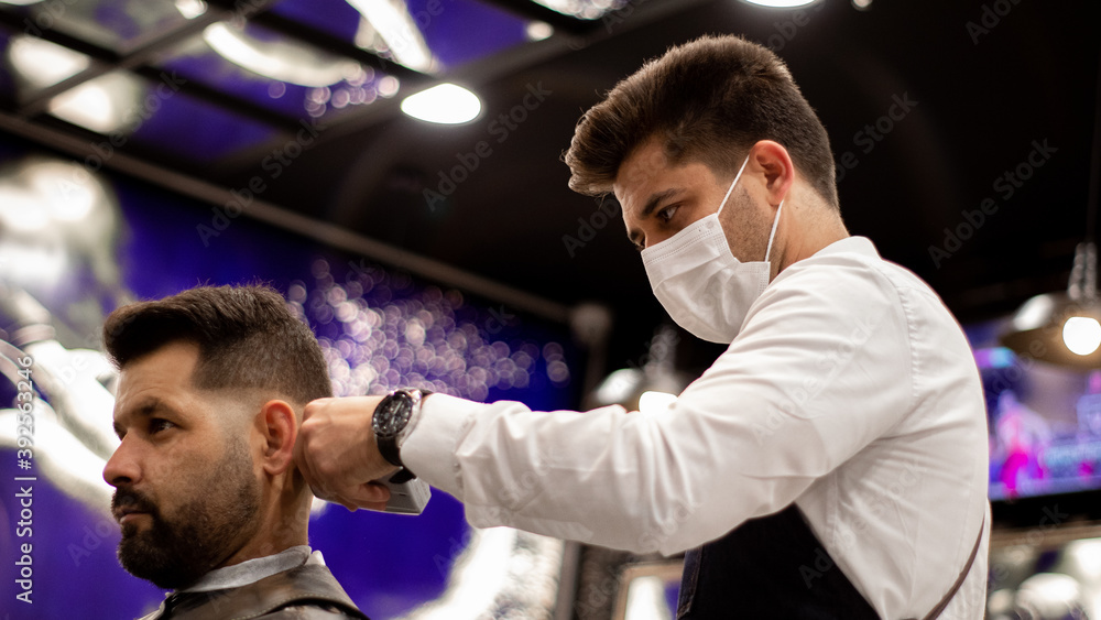 The young and elegant barber with mask cuts the hair of a client with Caucasian features The young and elegant barber with mask cuts the hair of a client with Caucasian features