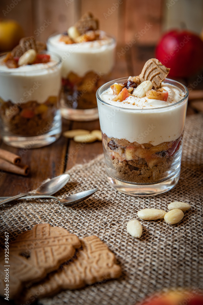 Kuchen im Glas mit Stücken vom Bratapfel, Zimt, Spekulatius, Apfel, Mandeln und Rosinen mit einer Creme aus Quark oder Mascarpone auf einem Tisch aus Holz in einer gemütlichen Küche zu Weihnachten Kuchen im Glas mit Stücken vom Bratapfel, Zimt, Spekulatius, Apfel, Mandeln und Rosinen mit einer Creme aus Quark oder Mascarpone auf einem Tisch aus Holz in einer gemütlichen Küche zu Weihnachten