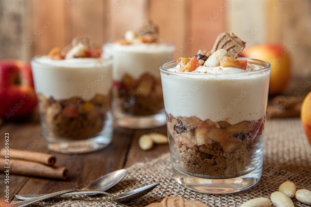 Kuchen im Glas mit Stücken vom Bratapfel, Zimt, Spekulatius, Apfel, Mandeln und Rosinen mit einer Creme aus Quark oder Mascarpone auf einem Tisch aus Holz in einer gemütlichen Küche zu Weihnachten Kuchen im Glas mit Stücken vom Bratapfel, Zimt, Spekulatius, Apfel, Mandeln und Rosinen mit einer Creme aus Quark oder Mascarpone auf einem Tisch aus Holz in einer gemütlichen Küche zu Weihnachten