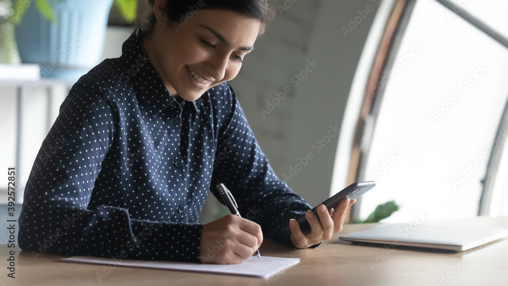 Close up smiling Indian woman holding phone, writing notes, sitting at desk, happy young female student intern watching webinar, online course, studying, preparing for exam, searching information Close up smiling Indian woman holding phone, writing notes, sitting at desk, happy young female student intern watching webinar, online course, studying, preparing for exam, searching information