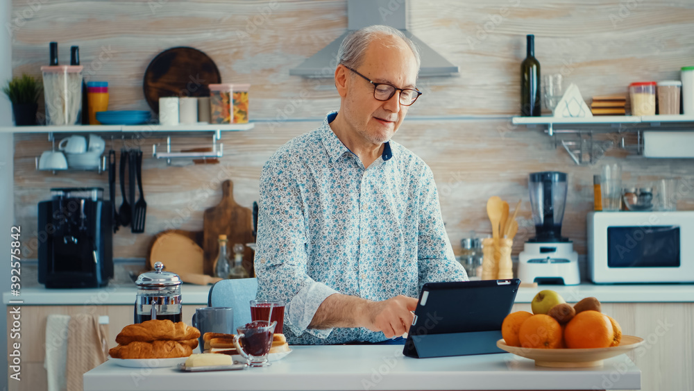 Senior parent browsing on tablet pc in kitchen during breakfast having a healthy lifestyle. Elderly person with tablet portable pad PC in retirement age using mobile apps, modern internet online Senior parent browsing on tablet pc in kitchen during breakfast having a healthy lifestyle. Elderly person with tablet portable pad PC in retirement age using mobile apps, modern internet online