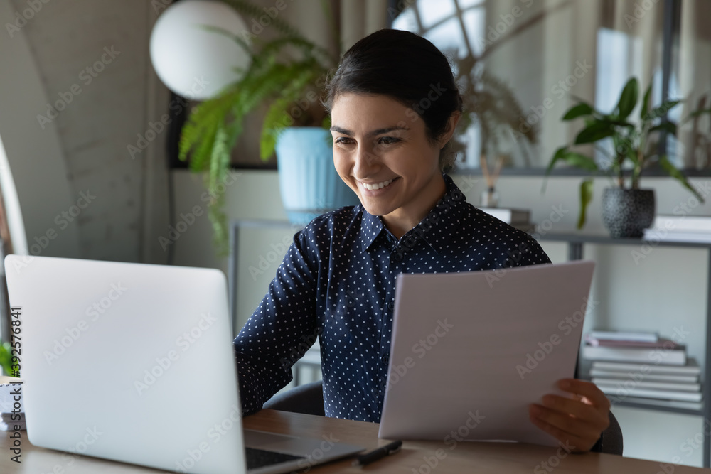 Smiling Indian businesswoman using laptop, checking financial documents, correspondence, happy employee holding paper sheet, looking at computer screen, satisfied by good news, great project results Smiling Indian businesswoman using laptop, checking financial documents, correspondence, happy employee holding paper sheet, looking at computer screen, satisfied by good news, great project results