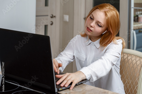 beautiful red-haired girl in a white blouse with a headset uses a laptop at home