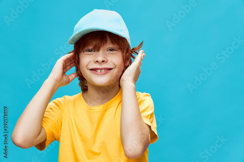 Smiling red-haired boy with a cap on his head holds his hands near the face yellow t-shirt