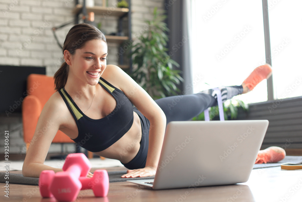 Fitness beautiful slim woman doing side plank with an elastic band and watching online tutorials on laptop, training in living room. Stay at home activities. Fitness beautiful slim woman doing side plank with an elastic band and watching online tutorials on laptop, training in living room. Stay at home activities.