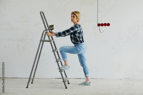 Young woman makes repairs in her house, next to a stepladder, holds a tool in her hands