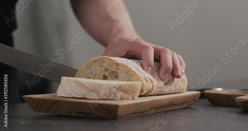 Slow motion man slicing ciabatta with bread knife on olive board