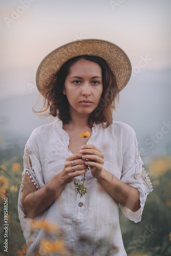 Obraz na płótnie brunette woman in a linen dress and straw hat walks among herbs and wildflowers on a meadow in the mountains. A woman in the Boho style, resting in the countryside, a simple village lifestyle w salonie