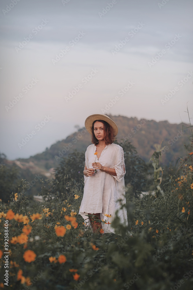 brunette woman in a linen dress and straw hat walks among herbs and wildflowers on a meadow in the mountains. A woman in the Boho style, resting in the countryside, a simple village lifestyle
