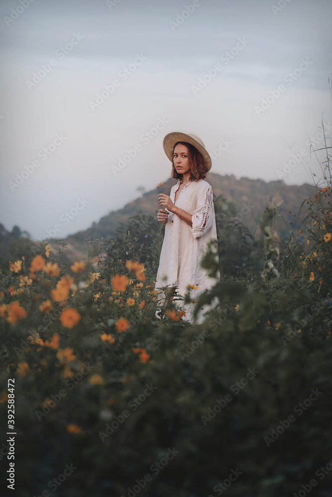 brunette woman in a linen dress and straw hat walks among herbs and wildflowers on a meadow in the mountains. A woman in the Boho style, resting in the countryside, a simple village lifestyle