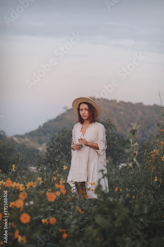 Obraz na płótnie brunette woman in a linen dress and straw hat walks among herbs and wildflowers on a meadow in the mountains. A woman in the Boho style, resting in the countryside, a simple village lifestyle w salonie