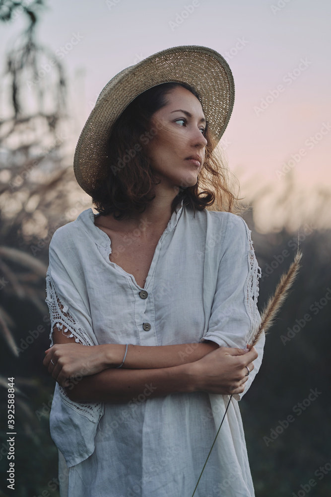 brunette woman in a linen dress and straw hat walks among herbs and wildflowers on a meadow in the mountains. A woman in the Boho style, resting in the countryside, a simple village lifestyle
