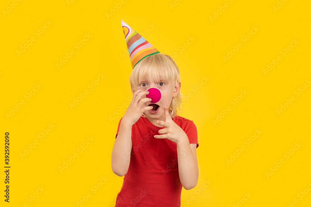 Surprised fair-haired boy with clown nose and party cap on yellow background. Kids party Surprised fair-haired boy with clown nose and party cap on yellow background. Kids party