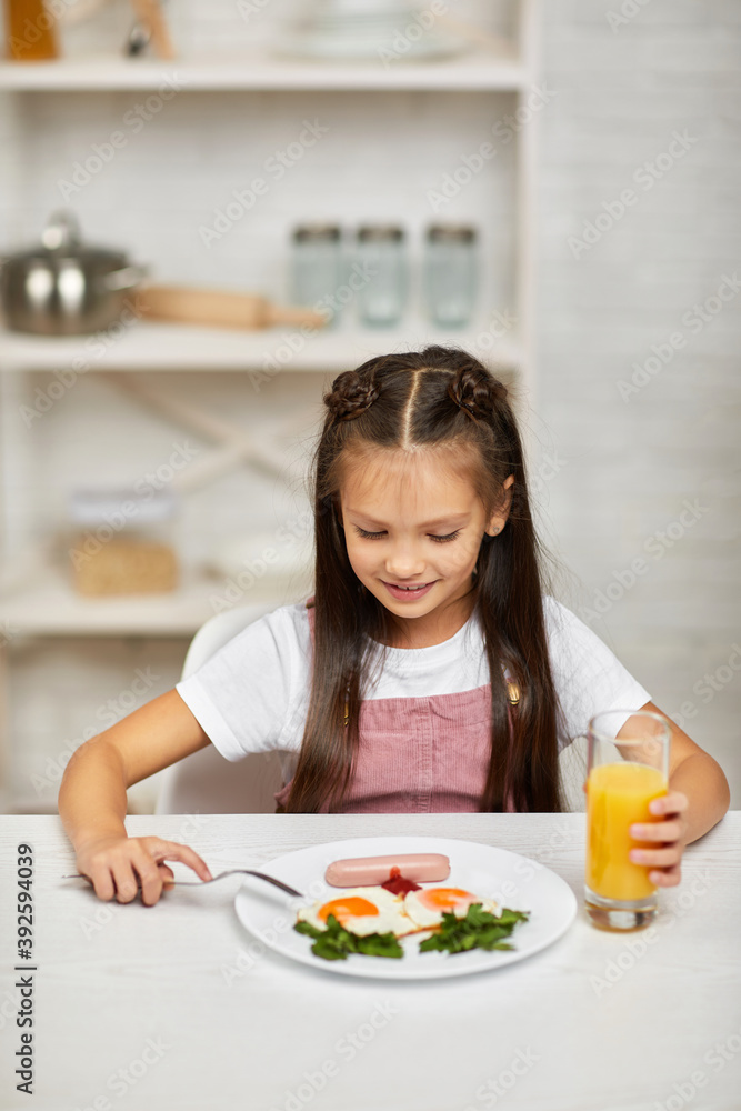 little child girl having breakfast - fried egg and orange juice in the kitchen. healthy breakfast. fried egg face smiles on a white plate