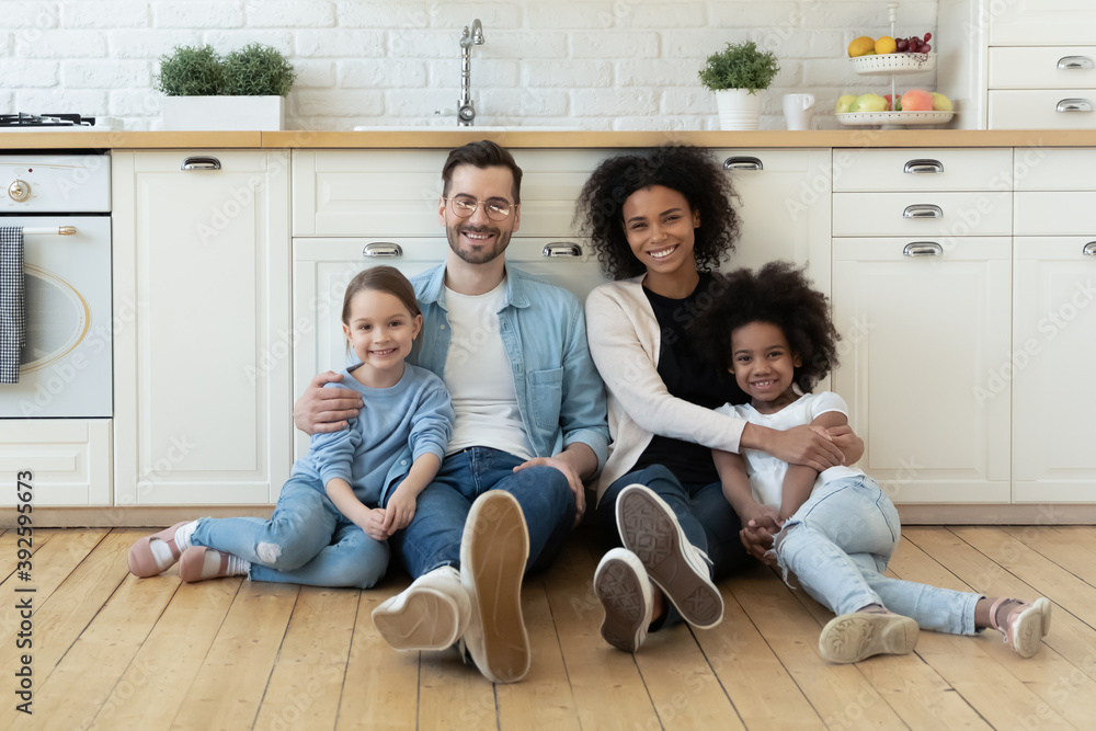 Portrait happy multiracial family sitting on warm wooden floor in modern kitchen at home, smiling Caucasian father and African American mother hugging two little daughters, posing for photo together - obrazy, fototapety, plakaty 