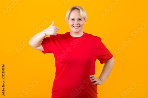 beautiful young plump female in red t-shirt on yellow background with hand gesture © Alexandr