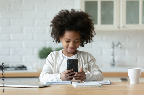 Pretty African American little girl using phone, distracted from studying, sitting at table with notebooks in modern kitchen, cute child holding smartphone, looking at screen, having fun