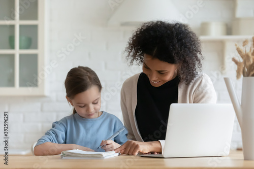 Smiling African American mother helping little adopted Caucasian daughter with school tasks  happy mum and pretty child schoolgirl sitting at table  doing homework  studying together  homeschooling