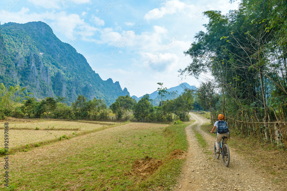 Woman riding mountain bike on dirt road in scenic landscape around Vang Vieng backpacker travel destination in Laos Asia rock pinnacles green valley