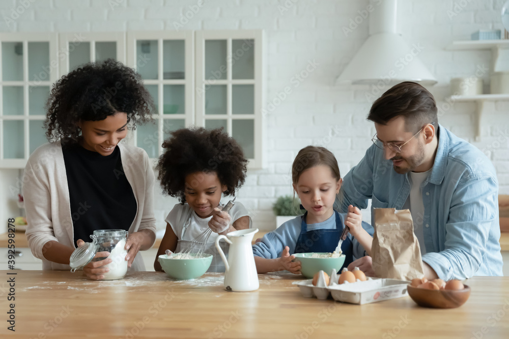 Happy diverse parents with two adorable little daughters wearing aprons cooking pancakes in kitchen, smiling African American mother and Caucasian father helping to kids, enjoying leisure time Happy diverse parents with two adorable little daughters wearing aprons cooking pancakes in kitchen, smiling African American mother and Caucasian father helping to kids, enjoying leisure time
