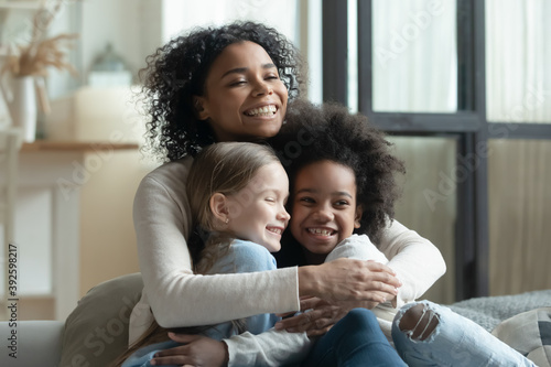 Overjoyed African American woman cuddling with two little daughters, multiracial family enjoying tender moment, sitting on cozy couch at home, loving mum hugging kids, adopted child photo