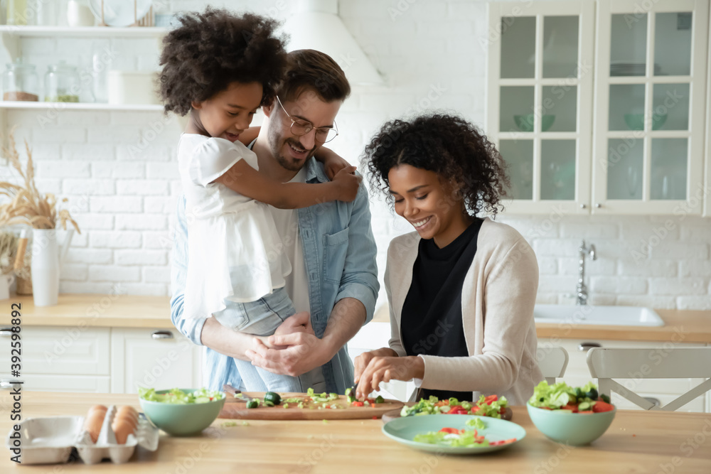 Caring father holding adorable little daughter, standing in kitchen, looking at African American mother cooking salad, shopping vegetables, multiracial family waiting for dinner or breakfast