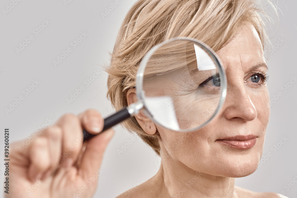 Close up portrait of attractive middle aged woman looking aside, holding a magnifying glass and showing her wrinkles, posing isolated over grey background Close up portrait of attractive middle aged woman looking aside, holding a magnifying glass and showing her wrinkles, posing isolated over grey background
