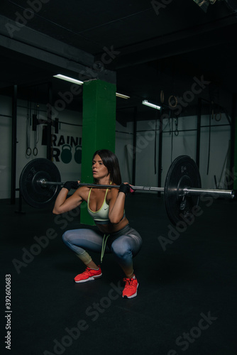 Fit young girl with light brown hair wearing black leggins, pink top and sneakers and doing squats with barbell, dark gym at background, cross training workout, portrait.
