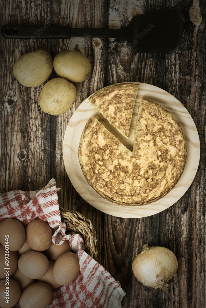 Aerial view of a potato omelette on a wooden board with ingredients: Eggs in a wicker basket, potatoes and onion Aerial view of a potato omelette on a wooden board with ingredients: Eggs in a wicker basket, potatoes and onion