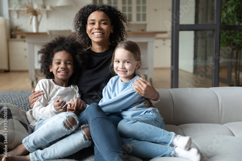Portrait of smiling African American mother hugging two little daughters, multiracial family concept, sitting on cozy couch in living room, posing for photo, looking at camera, adopted child