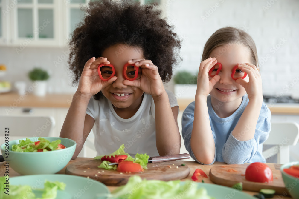 Close up head shot smiling diverse little girls holding red pepper as glasses, sitting at table with vegetables in kitchen, looking at camera, kids multiracial sisters posing for funny portrait Close up head shot smiling diverse little girls holding red pepper as glasses, sitting at table with vegetables in kitchen, looking at camera, kids multiracial sisters posing for funny portrait