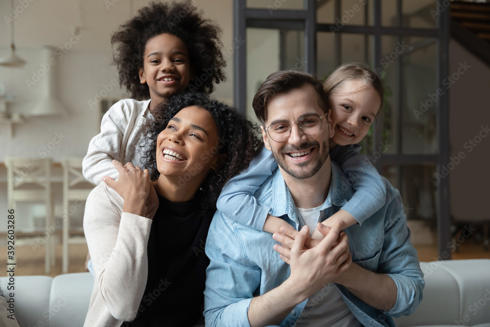 © fizkes - Head shot portrait happy multiracial family looking at camera, African American wife and Caucasian husband piggy backing two little daughters, posing for photo, having fun, sitting on couch