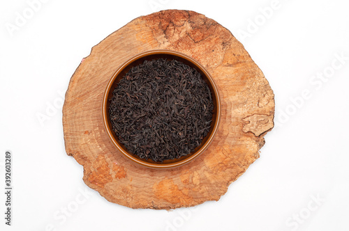 Dry tea leaves in brown ceramic bowl on wooden plate, isolated on white
