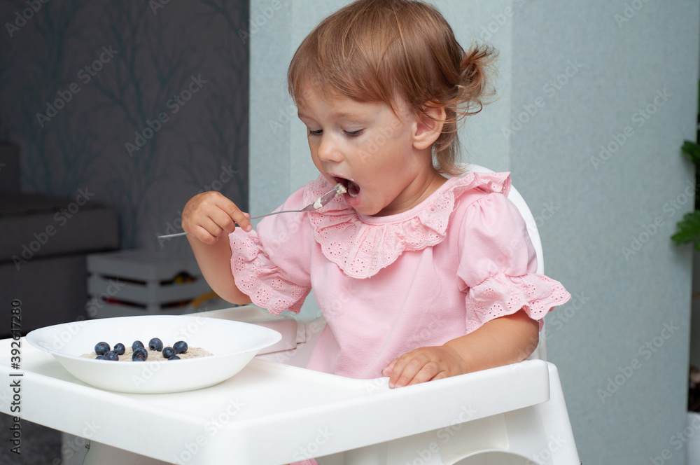Little kid girl eating her morning porridge oatmeal with blueberries herself, sitting in baby chair in kitchen Little kid girl eating her morning porridge oatmeal with blueberries herself, sitting in baby chair in kitchen