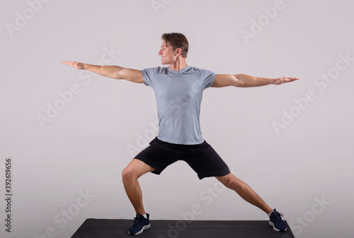 Handsome young man in sportswear doing warrior yoga pose on sports mat over light background, full length portrait photo