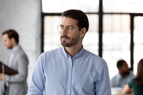 I like my job. Calm confident young businessman standing in large corporate office at workday looking aside with pensive smile feeling content happy, calling achievement pleasant work moment to mind