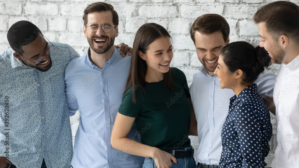 Obraz premium Sincere laughter. Overjoyed millennial multiethnic friends gathered together on informal meeting, young diverse colleagues taking short break in office work standing in group hugging talking laughing