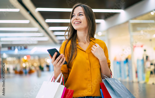 Happy beautiful young stylish woman with shopping bags is using smart phone while walking in the mall on black friday photo