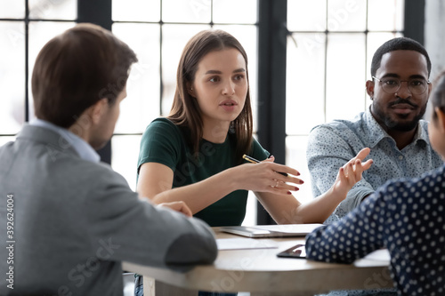 Two diverse multiethnic groups of business partners sitting at negotiation table, concentrated millennial female employee explaining plan project details to interested teammates on meeting in office photo