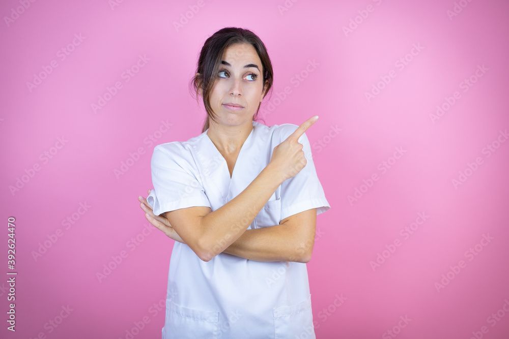 Young brunette doctor girl wearing nurse or surgeon uniform over isolated pink background smiling happy pointing with hand and finger to the side Young brunette doctor girl wearing nurse or surgeon uniform over isolated pink background smiling happy pointing with hand and finger to the side