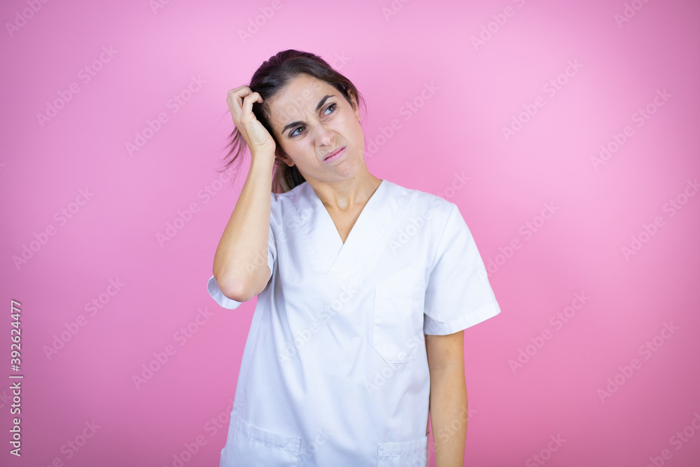 Young brunette doctor girl wearing nurse or surgeon uniform over isolated pink background confuse and wonder about question. Uncertain with doubt, thinking with hand on head Young brunette doctor girl wearing nurse or surgeon uniform over isolated pink background confuse and wonder about question. Uncertain with doubt, thinking with hand on head