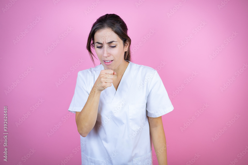 Young brunette doctor girl wearing nurse or surgeon uniform over isolated pink background with her hand to her mouth because she's coughing Young brunette doctor girl wearing nurse or surgeon uniform over isolated pink background with her hand to her mouth because she's coughing