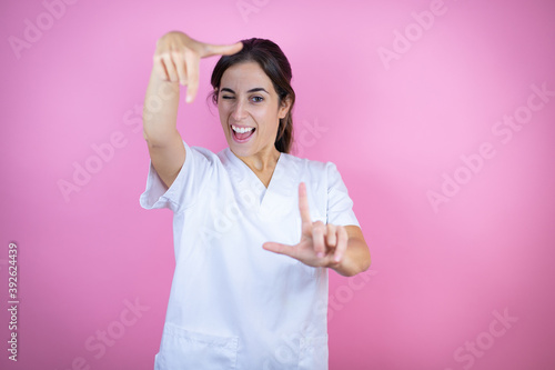Young brunette doctor girl wearing nurse or surgeon uniform over isolated pink background smiling making frame with hands and fingers with happy face