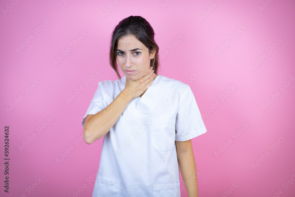 Young brunette doctor girl wearing nurse or surgeon uniform over isolated pink background touching painful neck, sore throat for flu, clod and infection Young brunette doctor girl wearing nurse or surgeon uniform over isolated pink background touching painful neck, sore throat for flu, clod and infection