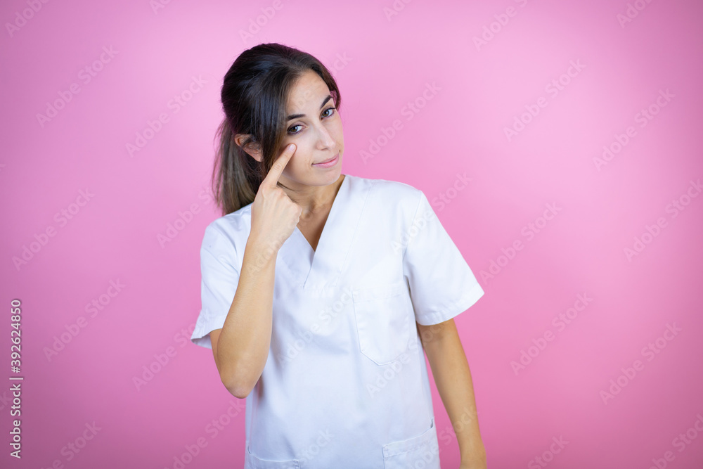 Young brunette doctor girl wearing nurse or surgeon uniform over isolated pink background Pointing to the eye watching you gesture, suspicious expression Young brunette doctor girl wearing nurse or surgeon uniform over isolated pink background Pointing to the eye watching you gesture, suspicious expression