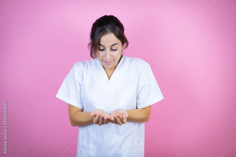Young brunette doctor girl wearing nurse or surgeon uniform over isolated pink background Smiling with hands palms together receiving or giving gesture. Hold and protection Young brunette doctor girl wearing nurse or surgeon uniform over isolated pink background Smiling with hands palms together receiving or giving gesture. Hold and protection